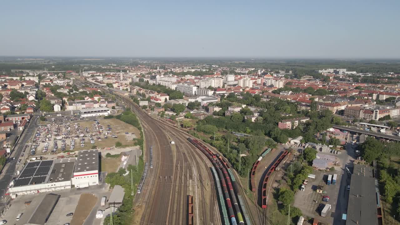 Aerial view of a city's railway hub with multiple train tracks converging near the urban center. Passenger and freight trains can be seen traveling and parked, surrounded by residential buildings.