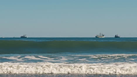 Waves-crashing-in-the-foreground-with-boats-in-the-background-on-the-blue-ocean-on-a-clear-day