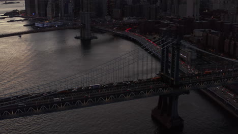 High-angle-view-of-slowly-moving-vehicles-on-large-suspension-bridge-over-river.-Tilt-up-reveal-of-downtown-skyscrapers-silhouettes-against-golden-sunset-sky.-Manhattan,-New-York-City,-USA