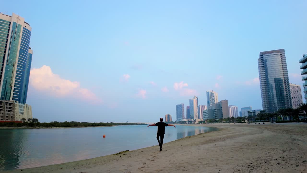 A silhouette of a man performs Tai Chi on an urban beach. Calm water, towering skyscrapers, and a soft sunrise create a peaceful, tranquil atmosphere