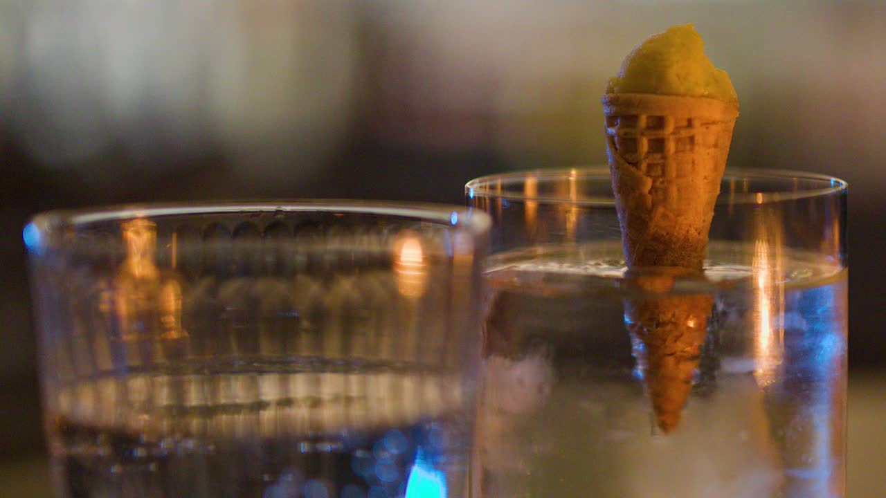 Close-up of cocktail glass with ice and cone garnish, warm evening bar lighting, shallow focus