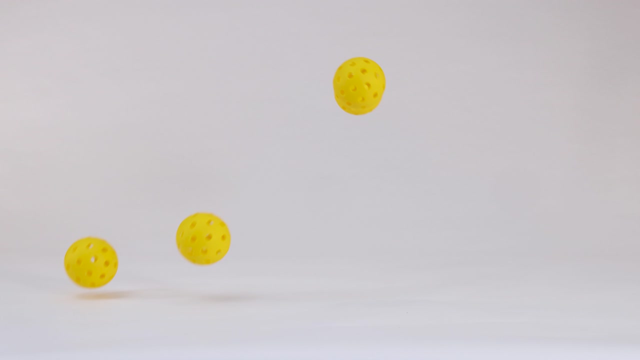 Three yellow plastic pickleball balls fall, bounce, and roll across a clean white studio floor