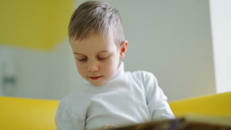 cute preschool boy indoors. portrait of a little boy sitting on sofa and looking down. child in white sweater reading a book at home.