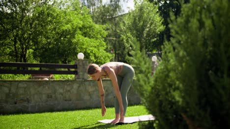 a blonde girl in special sports summer clothes does yoga on a gray mat on a green lawn in summer. sports activities in nature