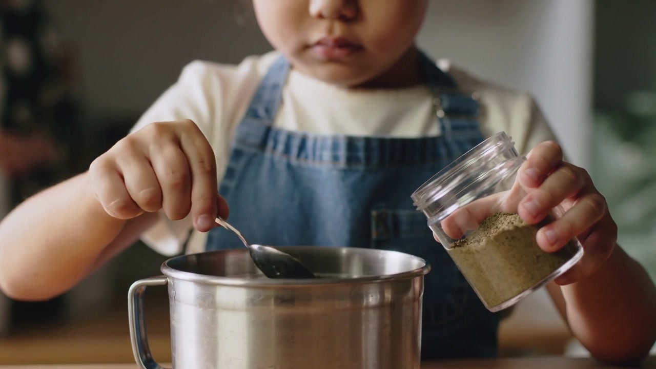 瓶とスプーンを持ったデニムエプロンの子供が、キッチンカウンターで緑の粉をボウルにすくい、料理をしている。