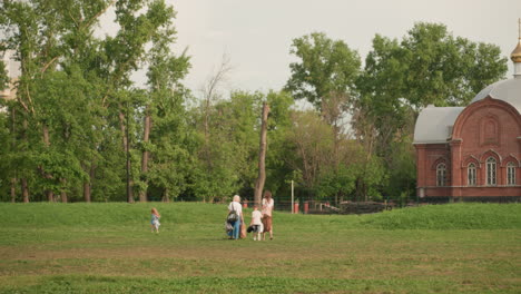 long shot of moms walking with kids across grassy park preparing for picnic, relaxed family outing, green trees background, casual summer clothes, children playing ahead, peaceful outdoor scene