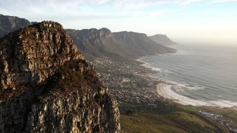 fly-by cinematic aerial shot of cape town's lions head peak revealing camps bay and table mountain national park during golden hour sunset