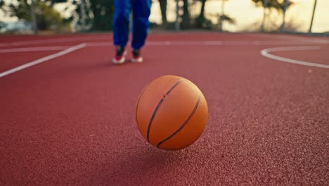 Shooting-close-up-a-blonde-girl-in-a-sports-uniform-picks-up-an-orange-basketball-from-the-red-floor-that-is-bouncing-on-a-street-court-in-the-morning