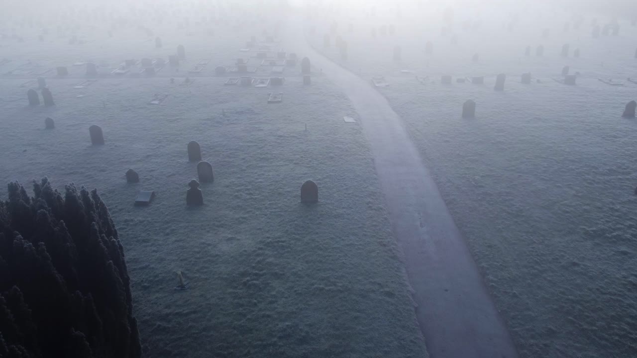 런던 도로의 안개가 자욱한 얼어붙은 세트퍼드 묘지 위로 공중 드론이 발사되어 경로 양쪽에 눈이 가득한 무덤 마당을 보여줍니다.