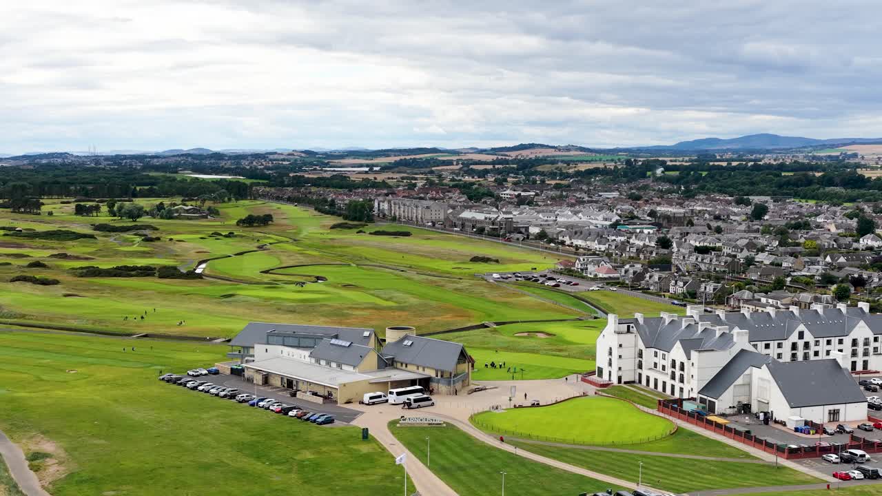 Drone glides over green fairways, sandy beach, and clubhouse under cloudy daylight in Dundee, Scotland