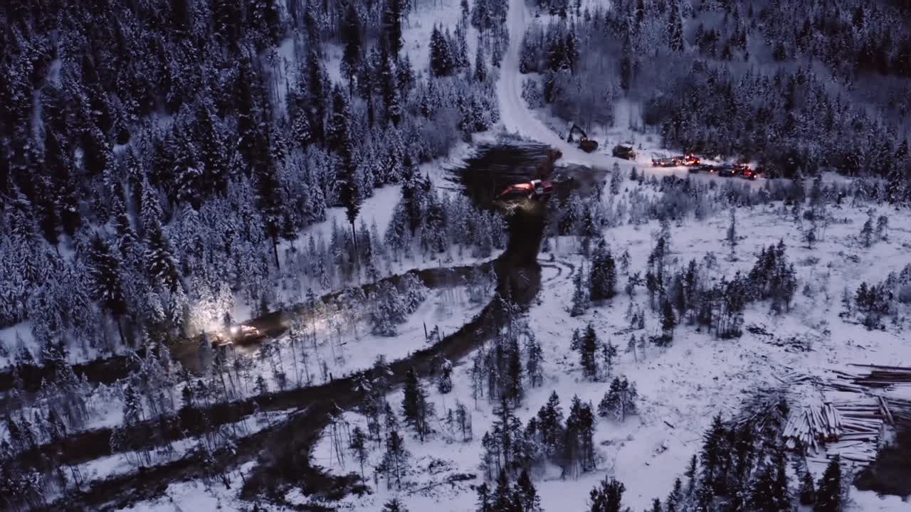 grúas de drones rápidamente en la escena del amanecer antes del amanecer donde 18 ruedas conducen a través de montones cubiertos de nieve de pinos maduros cortados en un campamento maderero