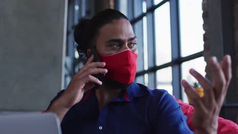 african american man wearing face mask sitting in cafe talking on smartphone using laptop