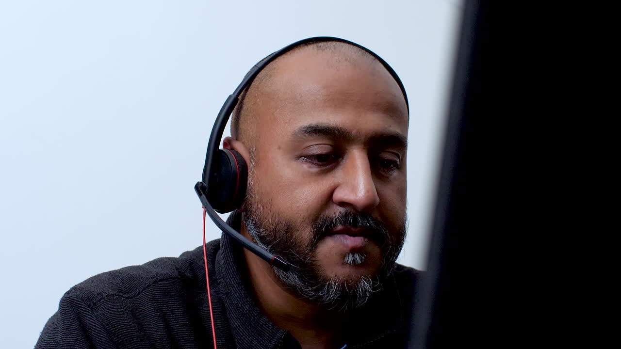 Close-up of a bald South Asian man wearing a headset, talking while working remotely from home