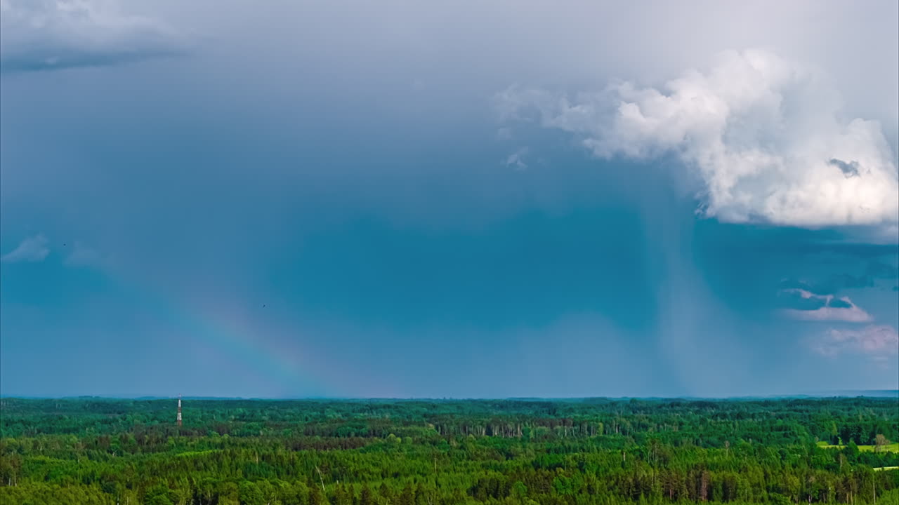 A beautiful aerial timelapse captures a faint rainbow appearing under dramatic rain clouds as they move across the sky above a vast and dense green forest
