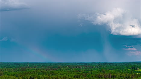 a beautiful aerial timelapse captures a faint rainbow appearing under dramatic rain clouds as they move across the sky above a vast and dense green forest