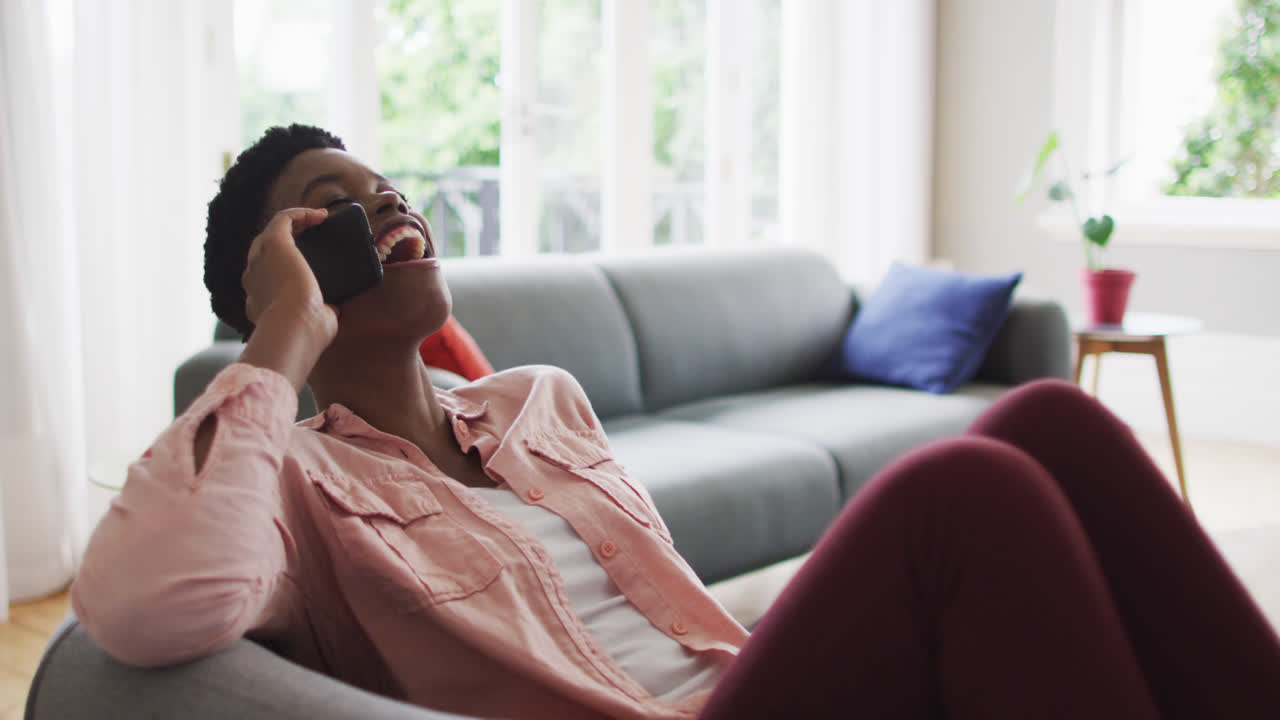 Afroamerikansk kvinde taler i smartphone, mens hun sidder på sofaen derhjemme