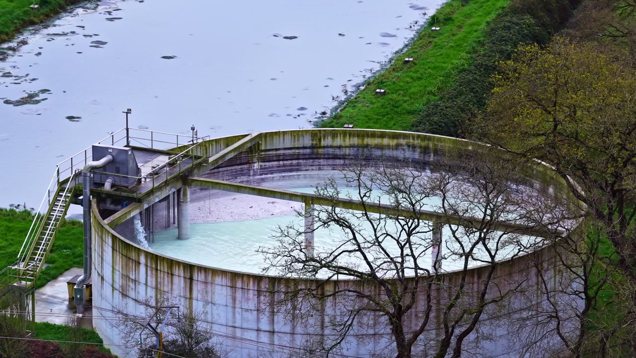 Aerial shot of a large circular industrial wastewater settling tank with metal walkways and stairs beside a muddy river surrounded by green grass and bare autumn trees on an overcast day