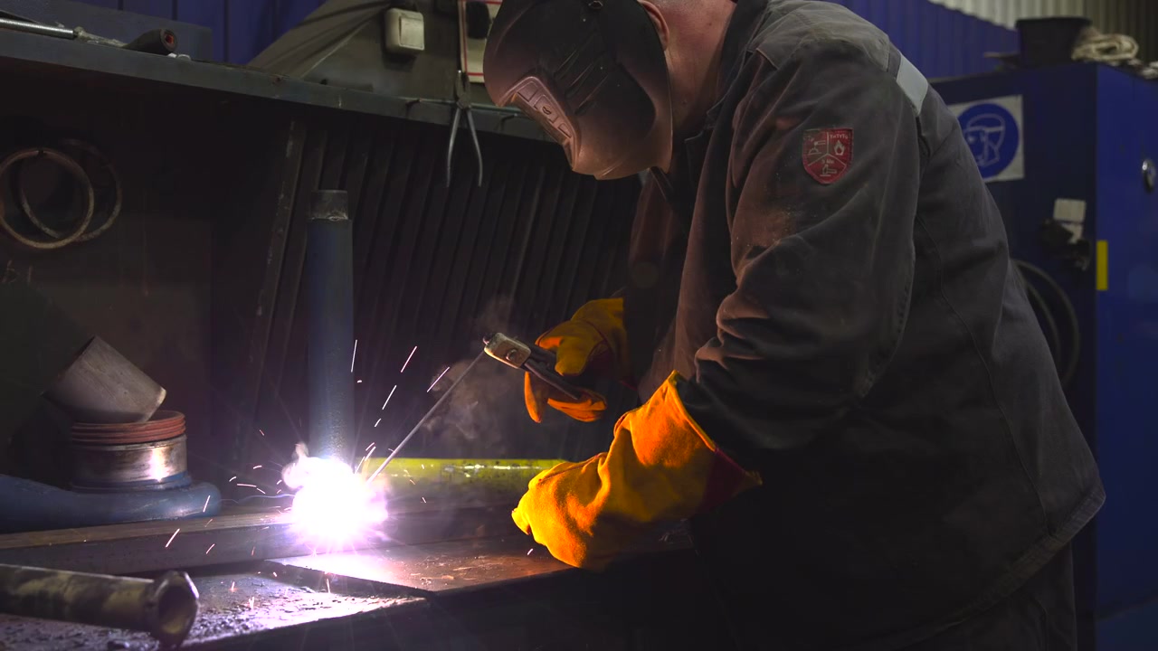 Protected worker in dark coverall and welding helmet bends over metal workbench, joining steel with vivid white arc, glowing sparks, rising smoke, and surrounding shop equipment in gritty industrial