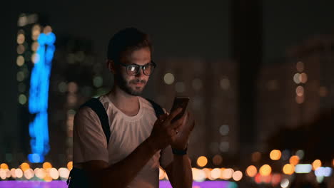 a young man with glasses at night looks at the smartphone screen and writes text messages for his blog on social networks. reads information and makes a trip martour