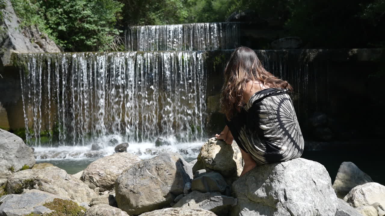 Young woman sitting on rocks beside waterfall in forest near Walensee, Switzerland, enjoying peaceful nature