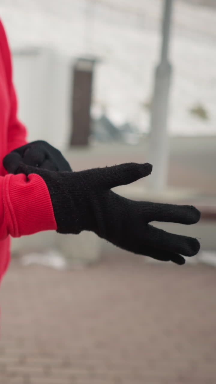 Woman putting on black winter glove for protection against cold outdoors, background shows modern benches and light pole, emphasizing winter urban landscape, dressed in cozy red hoodie