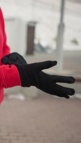 woman putting on black winter glove for protection against cold outdoors, background shows modern benches and light pole, emphasizing winter urban landscape, dressed in cozy red hoodie