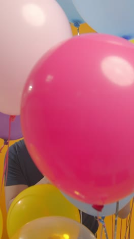 파티 모자를 입은 남자가 풍선과 파티 불꽃으로 생일을 축하하는 수직 비디오 스튜디오 초상화