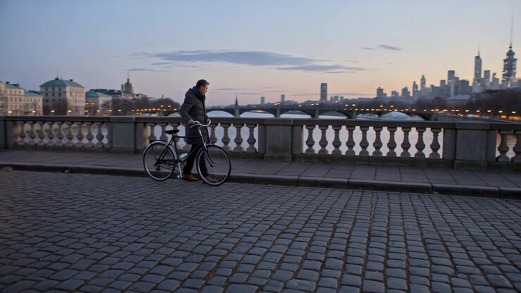 Un uomo con giacca cammina spingendo una bicicletta su un ponte fluviale al crepuscolo; una lampada lo fa fermare per ammirare lo skyline.