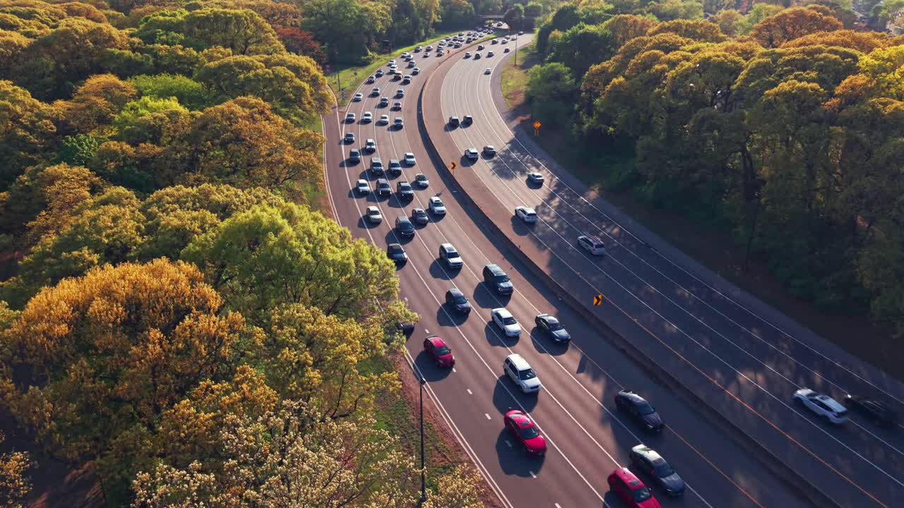An aerial view along the Southern State Parkway on Long Island, NY, taken during a bright and sunny day. The drone hovers next to the parkway, looking downward from above green and yellow treetops.