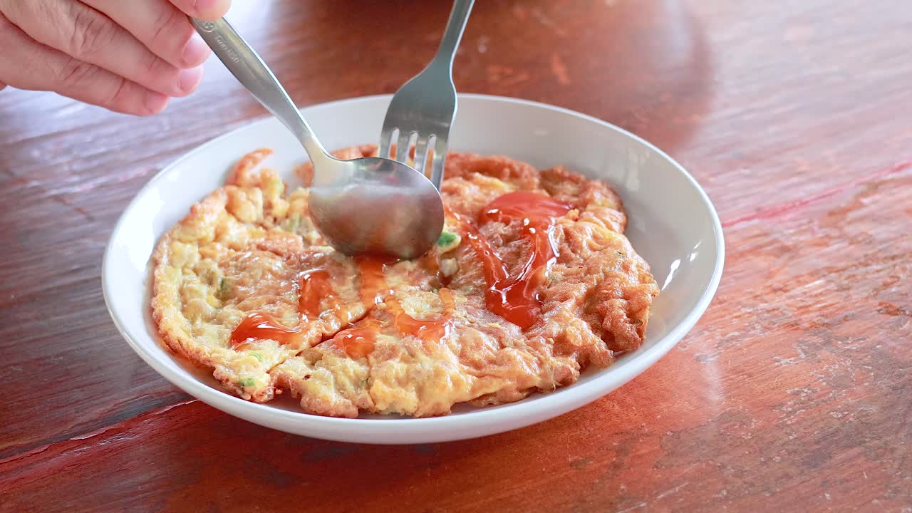A person enjoys a Thai-style omelette topped with chilli sauce, using a fork and spoon in a bright setting