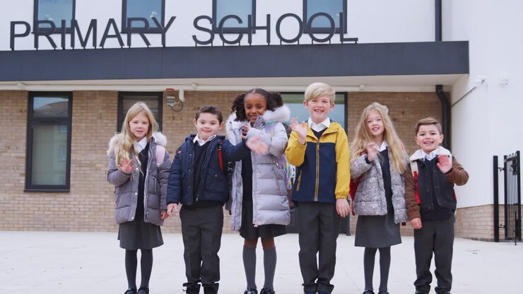 Primary school kids standing in front of their school looking to camera waving, front view
