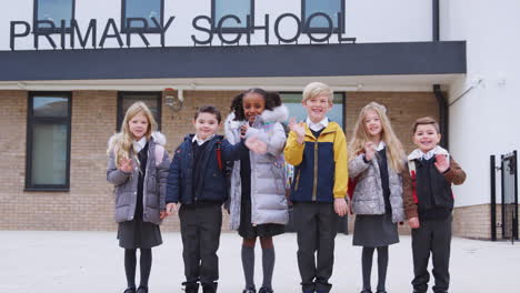 primary school kids standing in front of their school looking to camera waving, front view