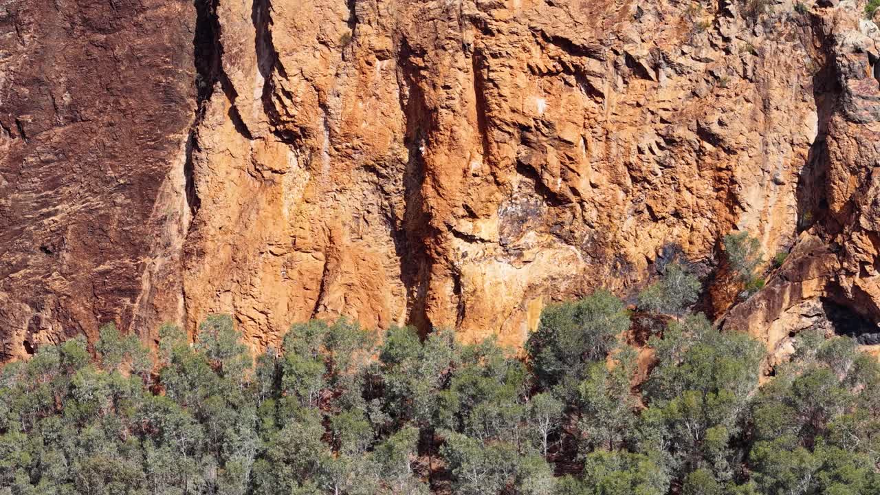 Aerial camera smoothly pans along rugged orange cliff face and dense green bushland in bright daylight, highlighting natural textures and Australian outback scenery
