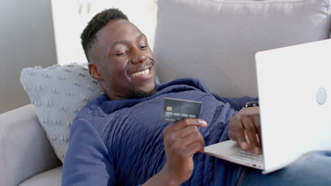 happy african american man lying and using laptop and credit card in sunny living room, slow motion