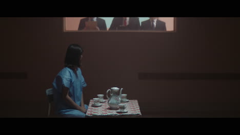 a girl sits at a table and strange figures in animal masks observes her from behind the glass