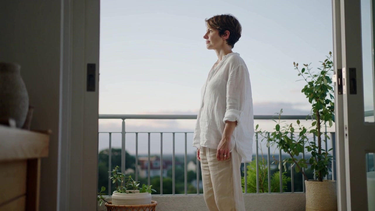 Donna in piedi con top di lino e pantaloni beige che guarda una porta scorrevole aperta, osservando le piante del balcone.