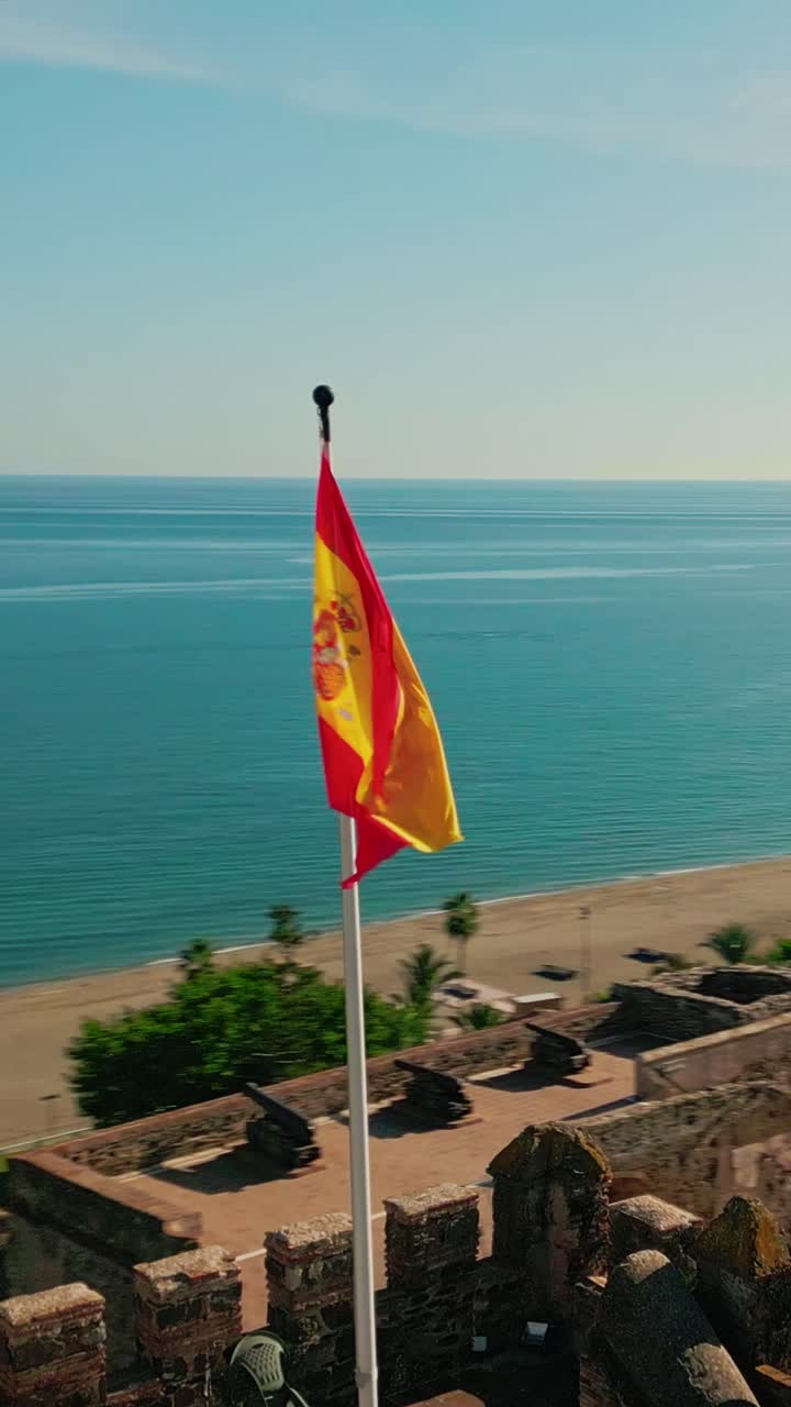 Vertikal luftbåren kredsløb af spansk flag, der vajer over historisk fæstning mod Middelhavskystlinjen, Sohail Castle, Fuengirola, Andalusien, Costa del Sol, Spanien.