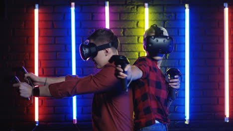 young man and woman in vr glasses and using joystick while playing a virtual reality game in a room with colorful neon lamps on the wall