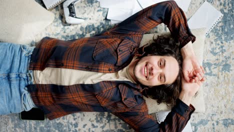 portrait of a happy brunette guy with curly hair in a checkered shirt who falls on a pillow on the floor and smiles posing among papers and a joystick in a modern apartment