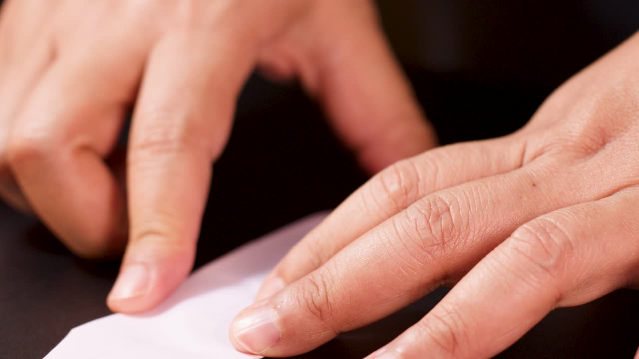 Close-up hands carefully folding pastel paper into an origami boat, warm lighting, static camera