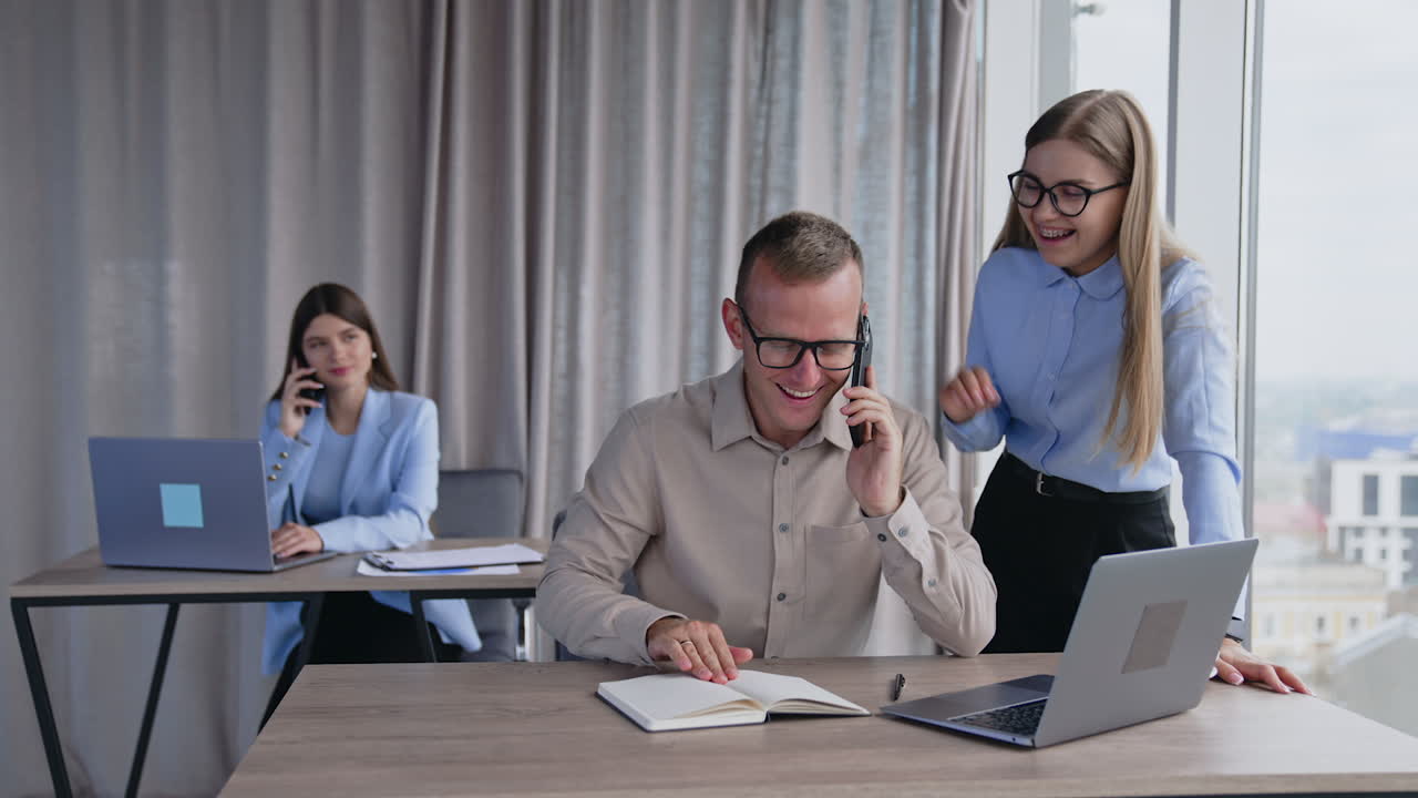 Smiling happy colleagues at the table on the foreground. Man speaks on the phone as well as woman at the desk behind.