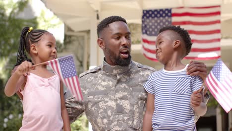 happy african american male soldier embracing his children with american flags, in slow motion