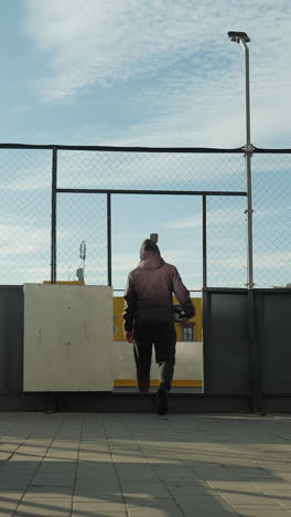 vista trasera de un individuo alejándose de la arena deportiva urbana, cargando una pelota de fútbol, con vibrantes contenedores de almacenamiento amarillos y edificios de la ciudad en el fondo, la luz del sol creando un resplandor