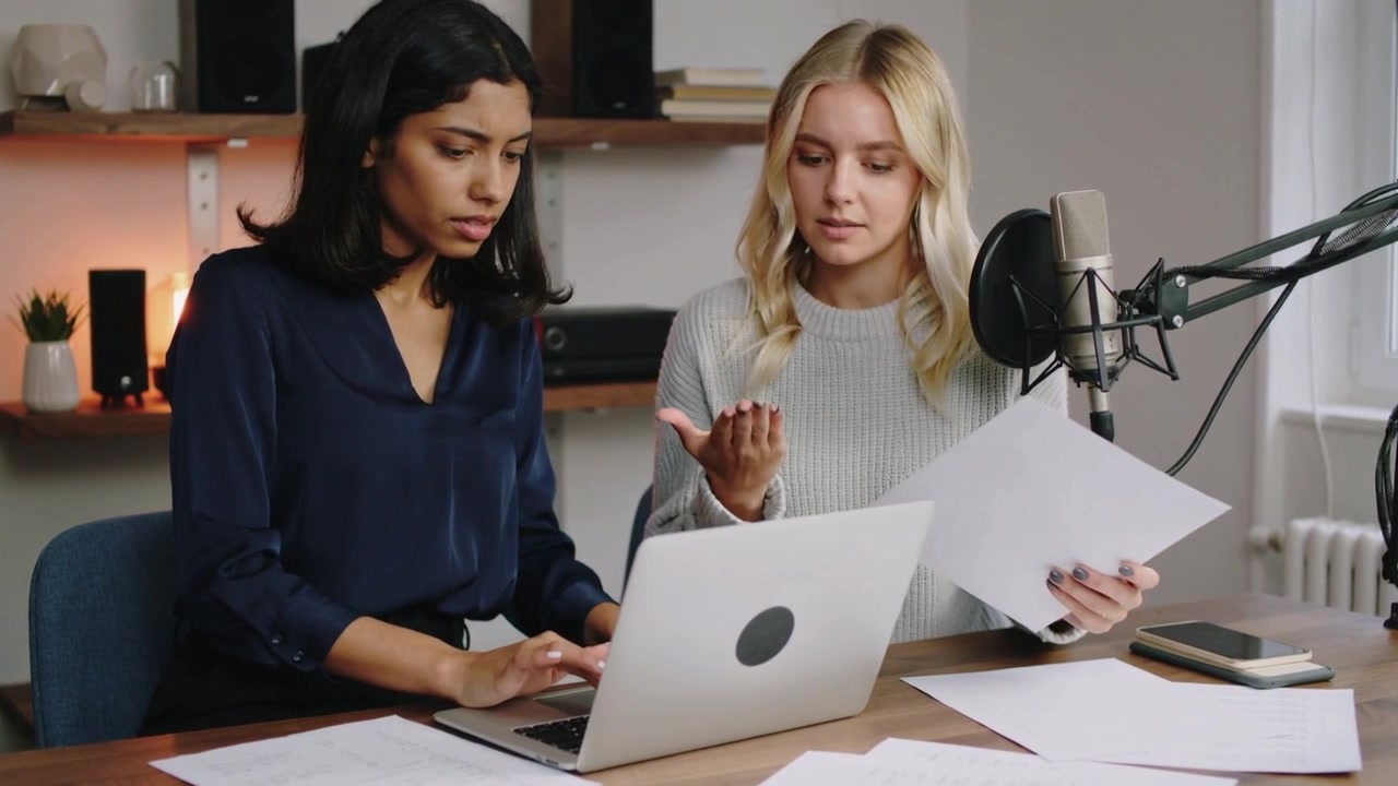 Sitting cohosts in navy blouse, typing on laptop and arranging papers near mic in podcast room