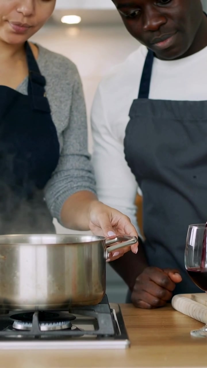Vertical video: Stirring cook in apron holding pot on lit stove, partner pouring wine for dinner