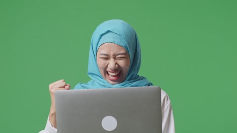 close up of asian muslim woman looking at a laptop then screaming goal celebrating while standing in the green screen background studio