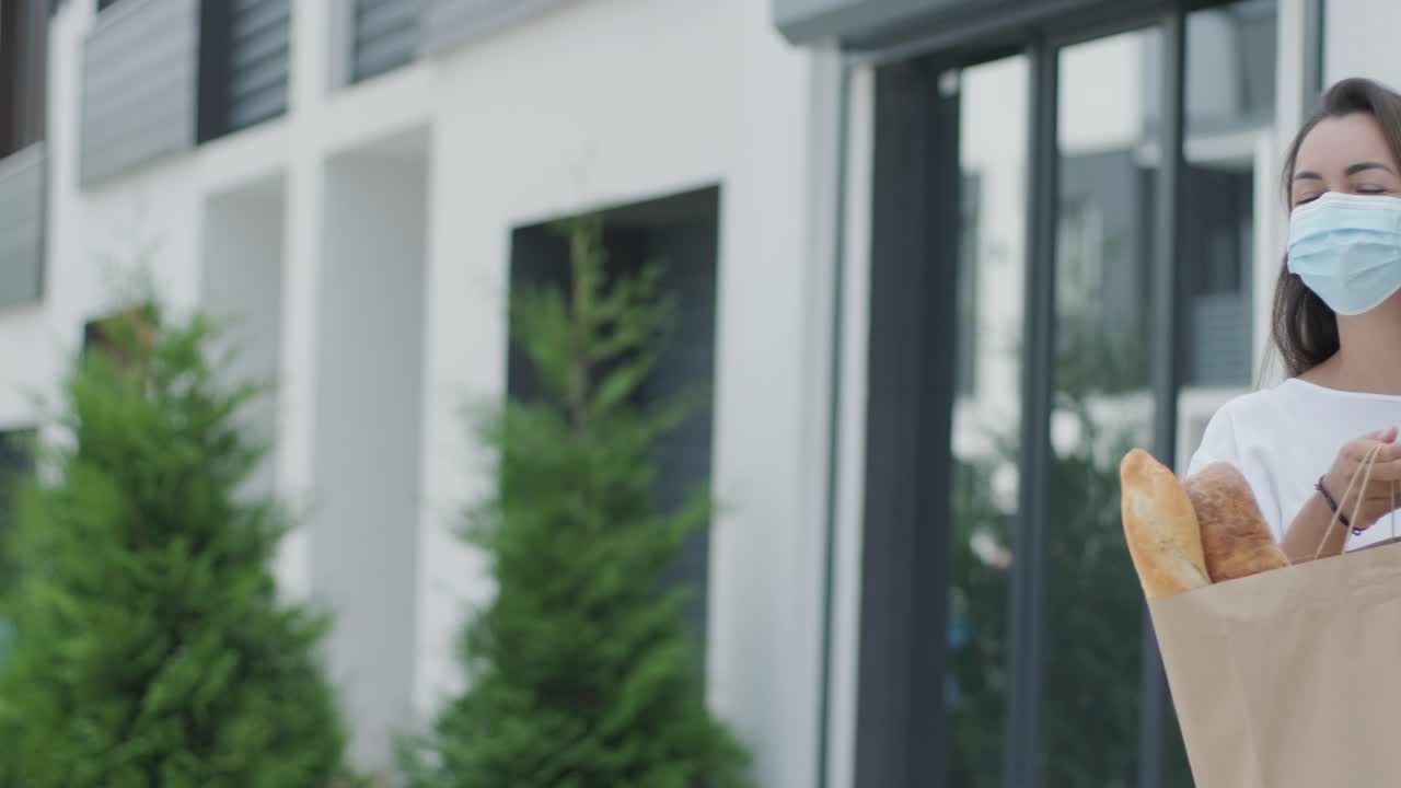 보호 마스크를 착용 한 아름다운 여성은 마스크와 장갑을 착용 한 배달자를 만나 그녀의 집 에 안전한 음식과 함께 쇼핑 패키지를 제공합니다.