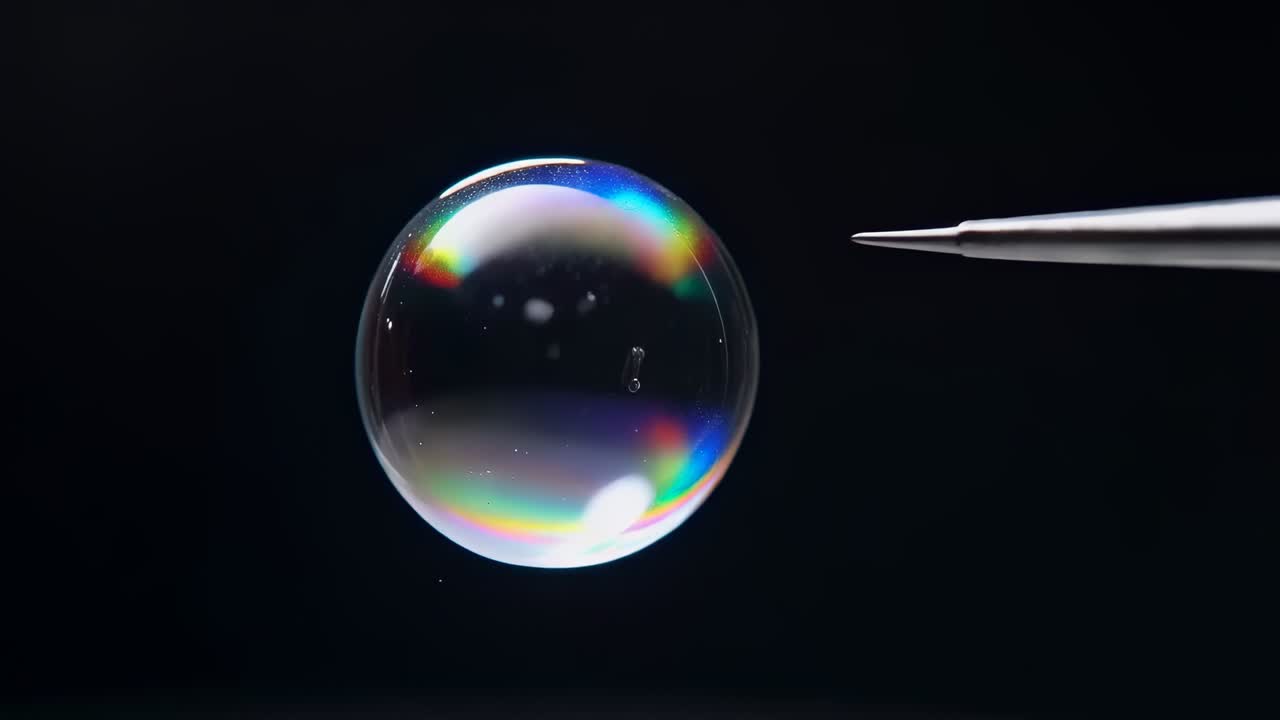 Metallisk spids der prikker hul i regnbuefarvet sæbeboble i et studie, mens dråber sprøjter og skaber bokeh