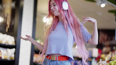 Happy-girl-with-pink-hair-in-a-purple-T-shirt-wearing-white-headphones-listens-to-music-and-dances-in-a-supermarket