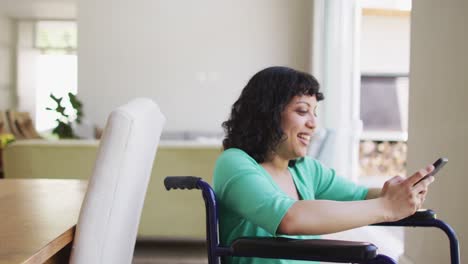 Happy-biracial-disabled-woman-in-wheelchair-using-smartphone-in-living-room